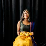 A South Asian woman in traditional attire holding Diwali sweets, smiling.